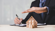 Business man in suit holding hand over small wood house with three small wood people.